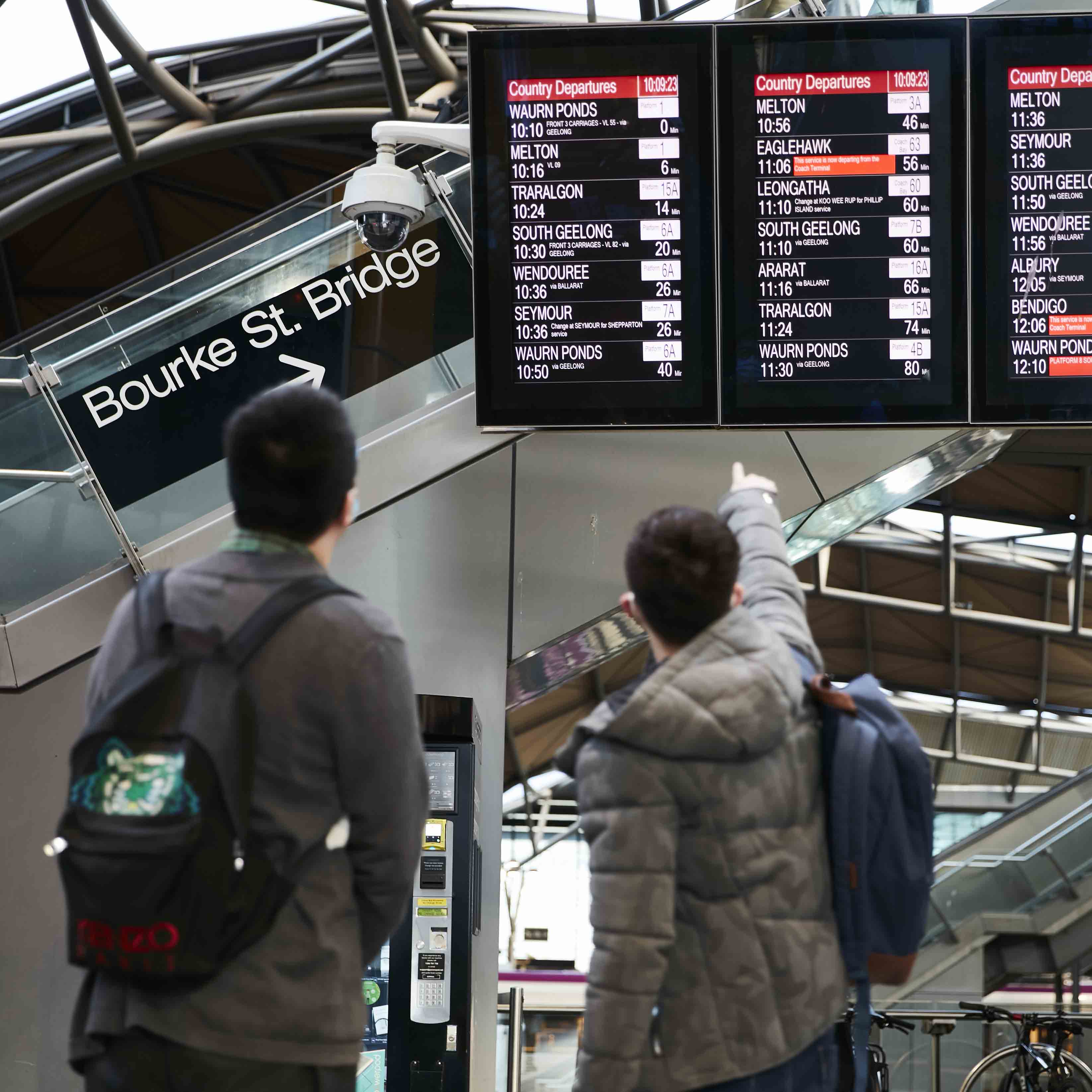 Image is a photograph of two people looking at the train departure displays at Southern Cross Station. The person on the right is pointing at the displays.