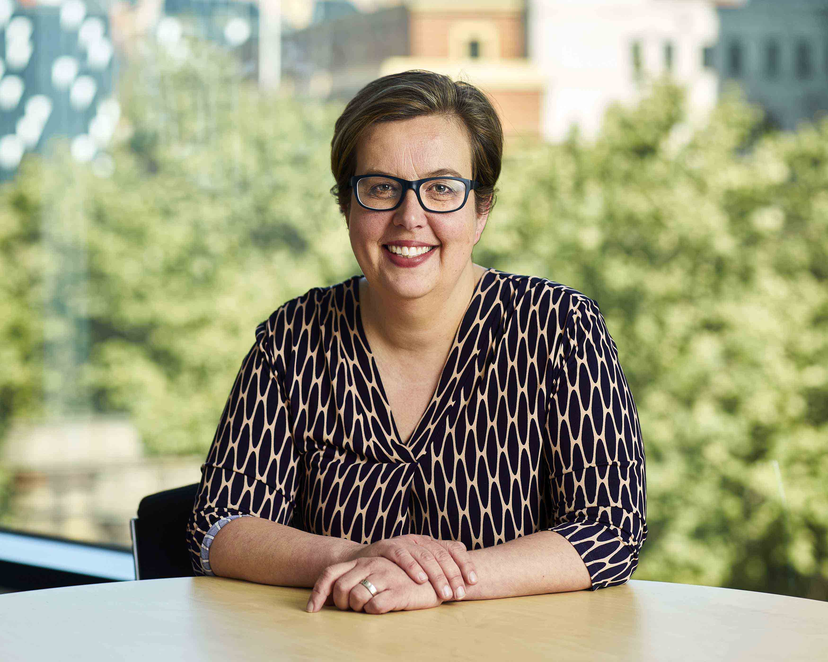 Image shows Acting Public Transport Ombudsman, Ann Jorgensen. Ann is a Caucasian woman with short-length brown hair. She is wearing a black and tan patterned dress and dark framed glasses. She is sitting at a table and looking directly at the camera. The blurred background shows trees and building in the Melbourne CBD.