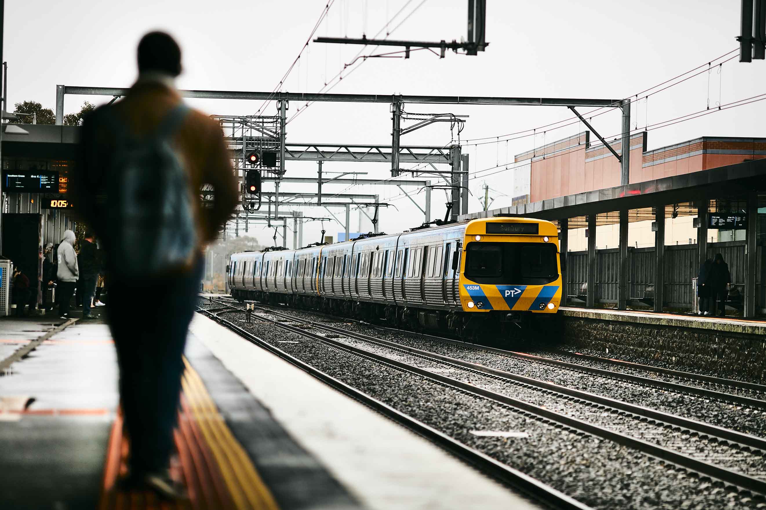 Image shows a person walking on a station platform in the foreground. In the background is a train on the Sunbury line as it pulls into a station. 