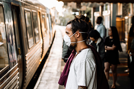Image shows a man standing on a train platform wearing a face mask during the COVID-19 pandemic while waiting for a train.