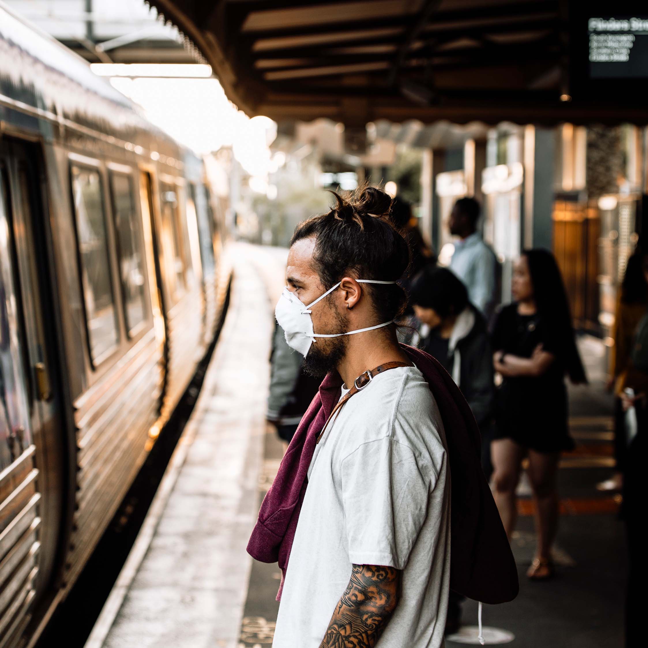 Image shows a man standing on a train platform wearing a face mask during the COVID-19 pandemic while waiting for a train.