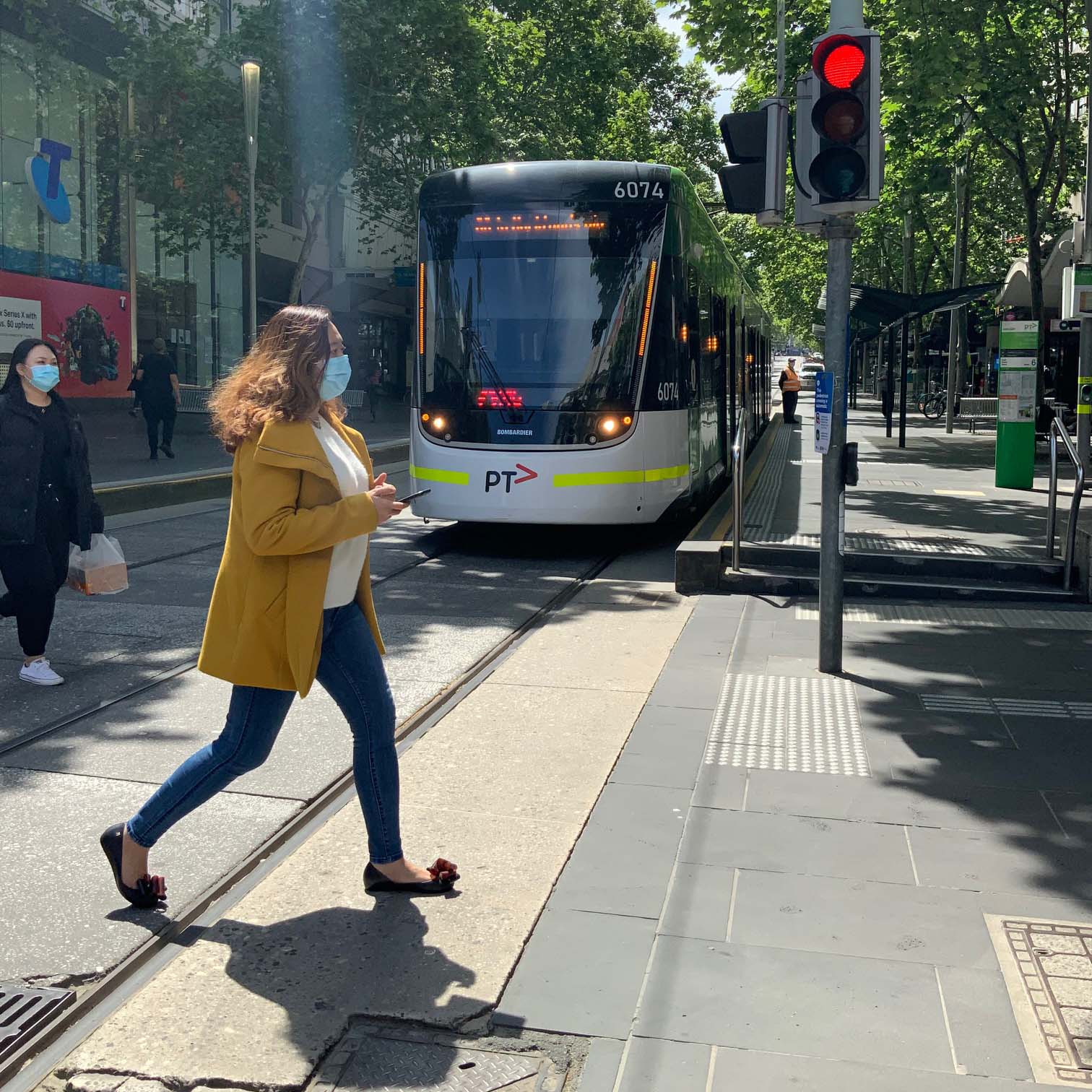 Image shows people walking across tram tracks while wearing face masks during the COVID-19 pandemic. There is a tram in the background.