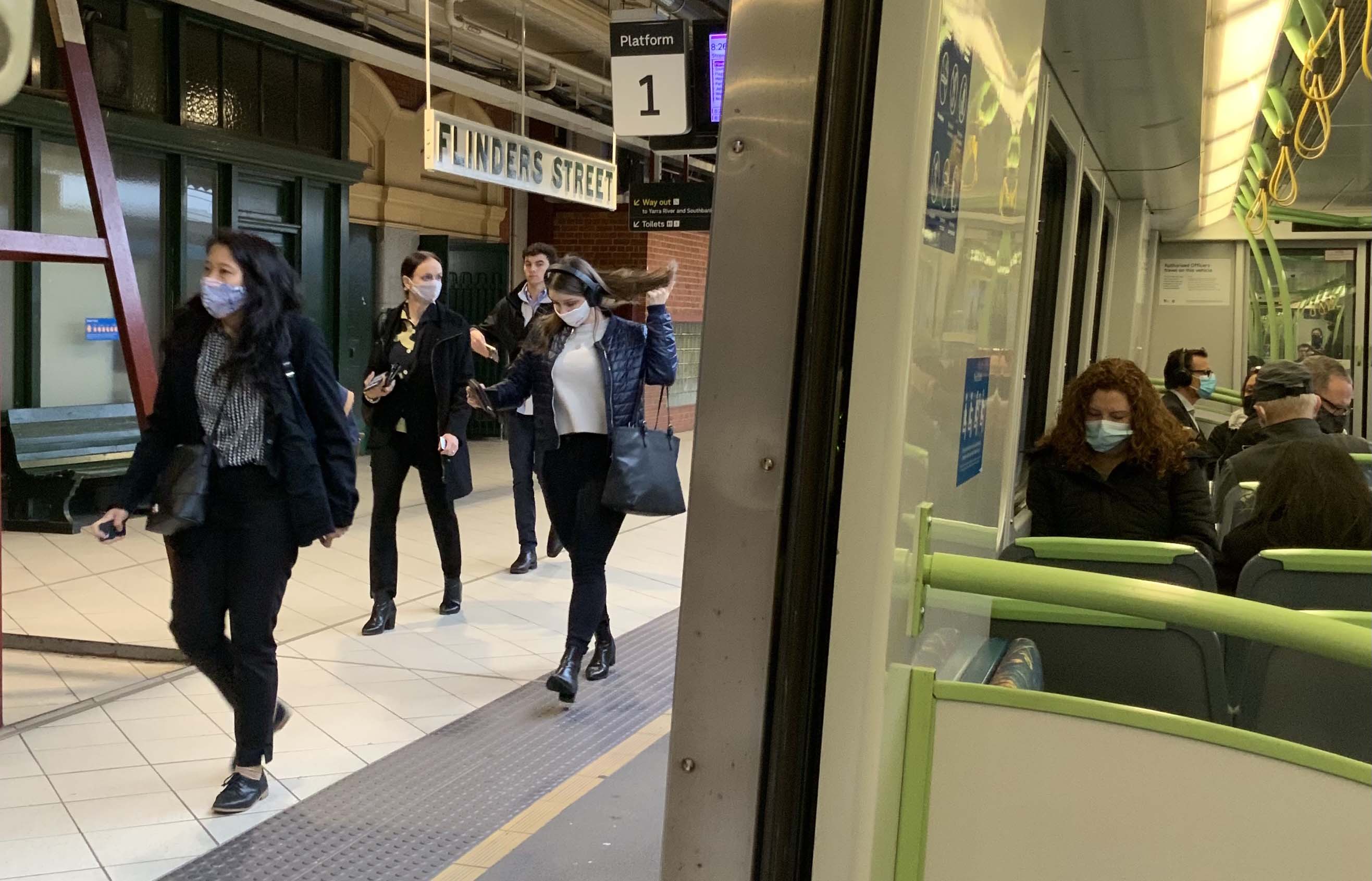 Image shows people wearing face masks whilst using public transport during the COVID-19 pandemic. The photo has been taken from inside a train and shows five people sitting on the train while wearing face masks. Also visible through the open train door are four people walking on the station platform past signs for Flinders Street Station and Platform 1.