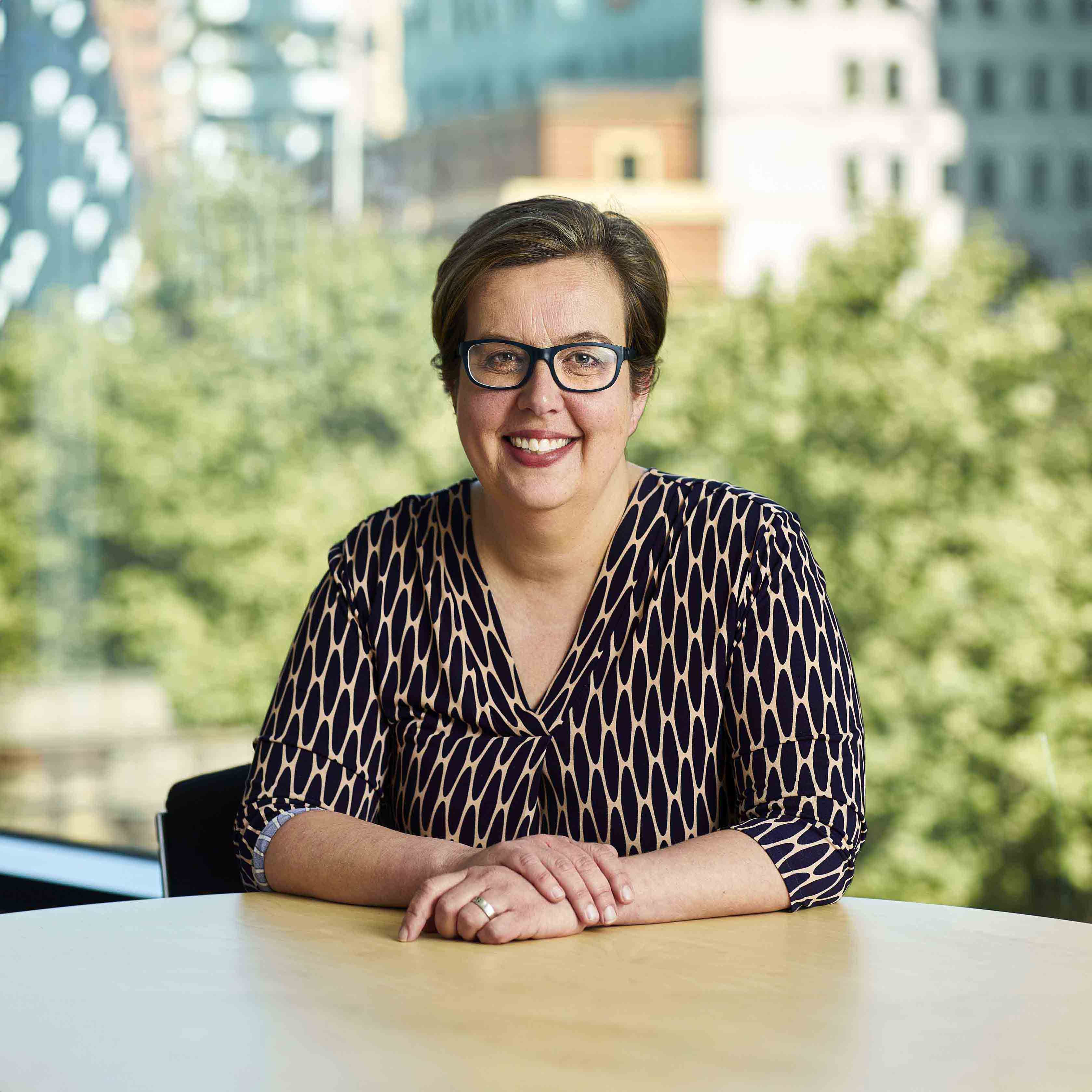 Public Transport Ombudsman Ann Jorgensen is a Caucasian woman. She has short brown hair and is wearing dark-framed glasses and a black and tan patterned dress. She is sitting at a table with her hands crossed before her and is looking directly into the camera and smiling. In the background are trees and a blurred cityscape.