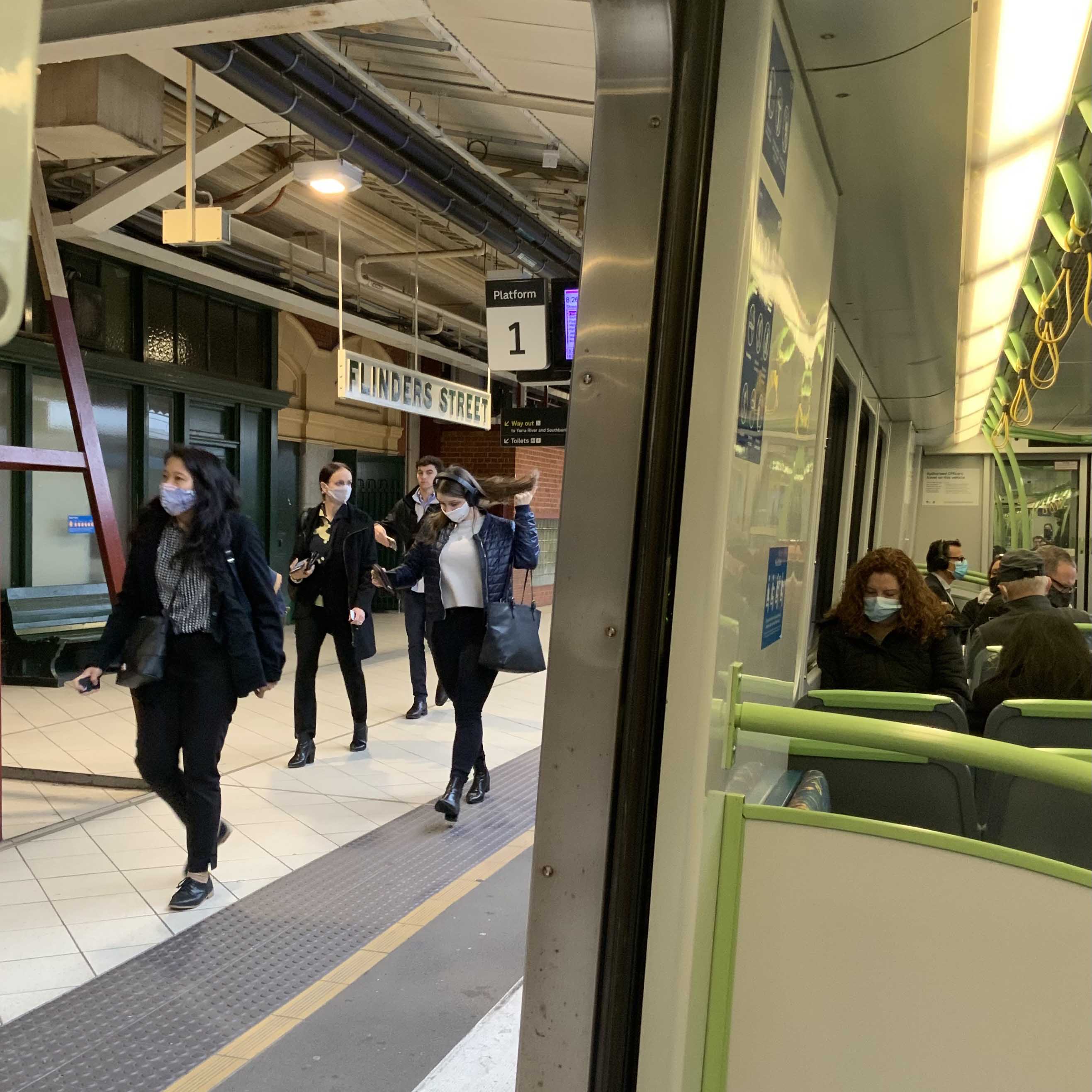 Image shows people wearing face masks whilst using public transport during the COVID-19 pandemic. The photo has been taken from inside a train and shows five people sitting on the train while wearing face masks. Also visible through the open train door are four people walking on the station platform past signs for Flinders Street Station and Platform 1.