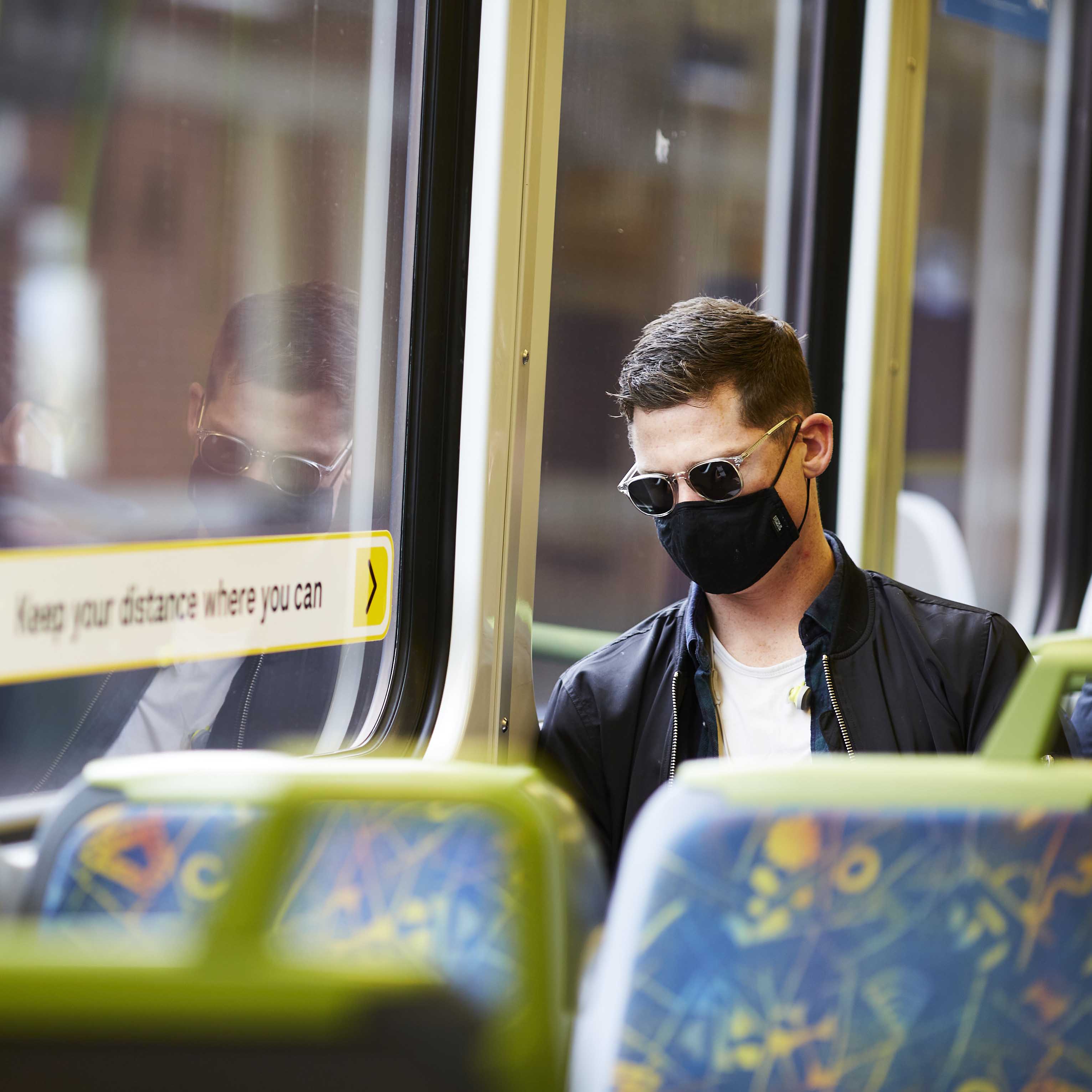 Image is a photograph of a passenger wearing a face mask while travelling on a Metro train. The passenger is a young Caucasian male with short brown hair. He is wearing dark sunglasses with clear frames, a black face mask, black leather jacket and a white t-shirt. He is looking down at his phone and leaning against the window of the train carriage. A blurred station platform is visible through the window.