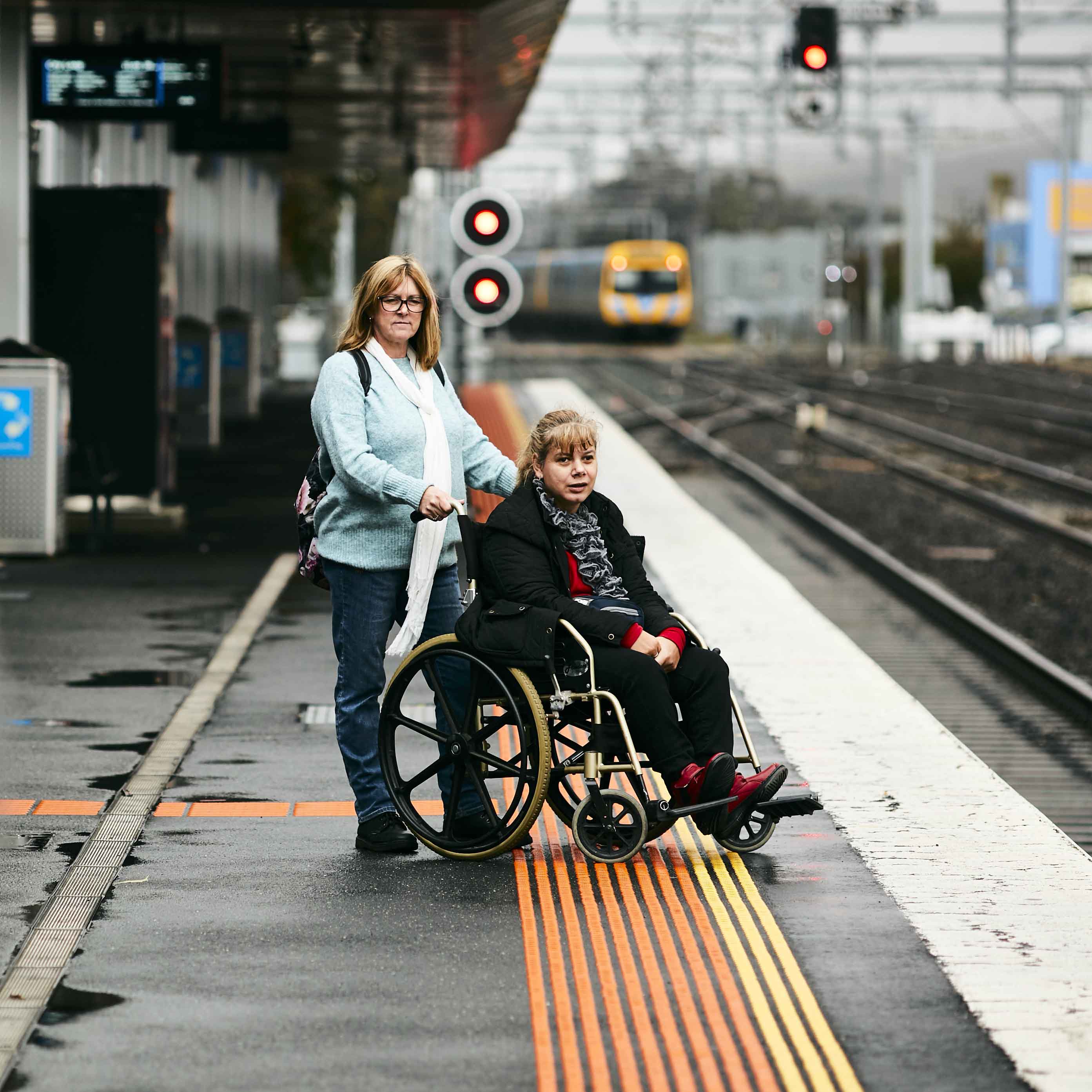 Image shows a person in a wheelchair with their carer waiting on a train station platform.