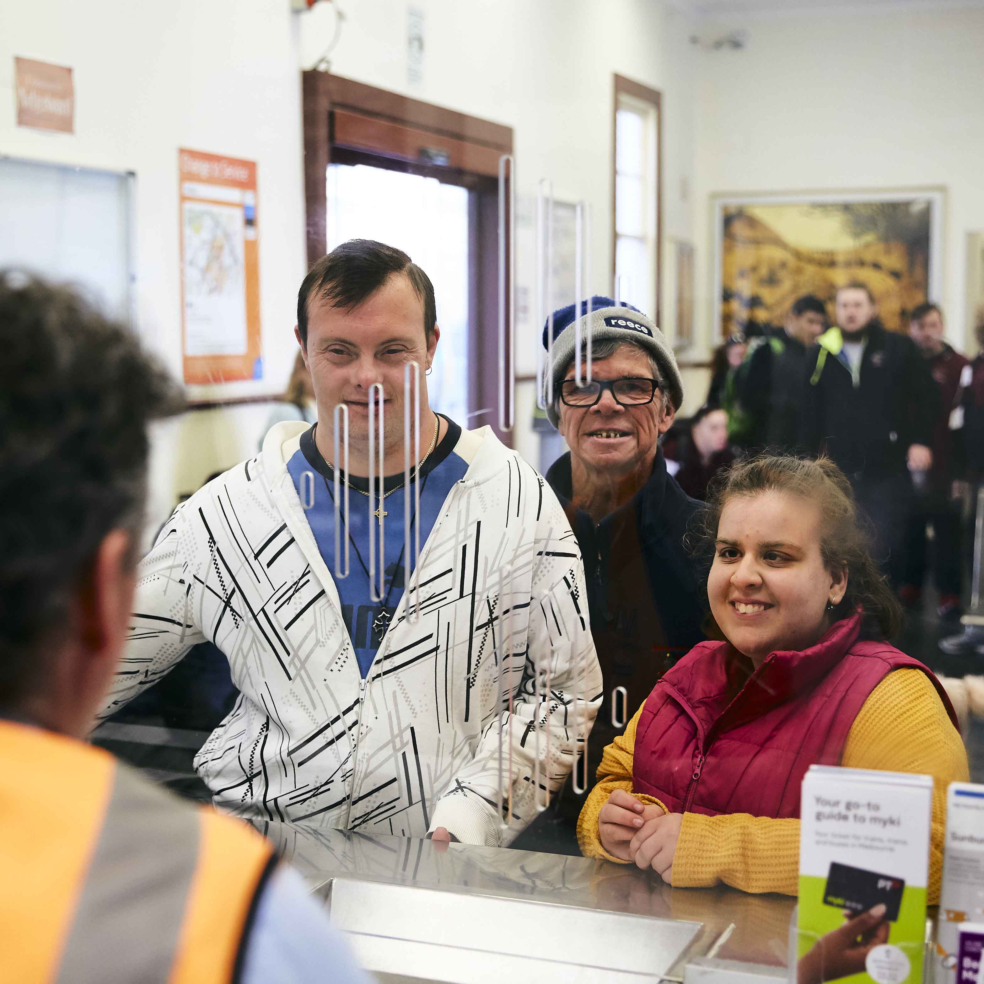 Image is a photograph of three people with disabilities standing at the service desk of a train station.