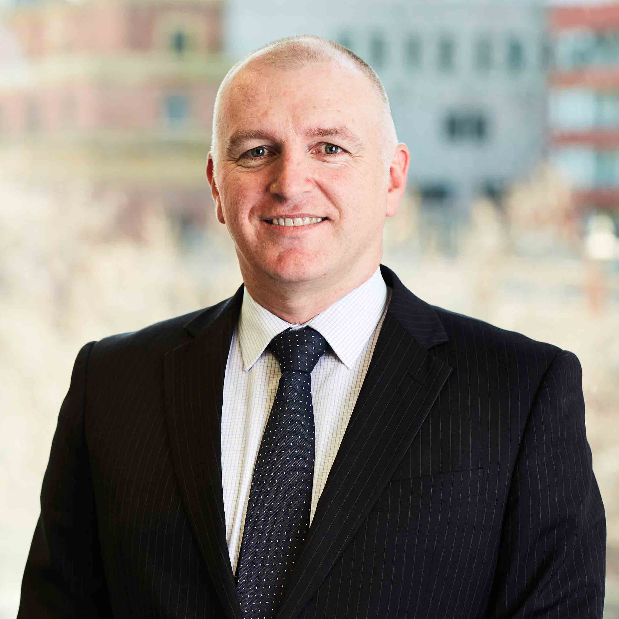 Image shows Public Transport Ombudsman, Simon McKenzie. Simon is a Caucasian man with closely cropped hair and he is smiling for the photo. He is wearing a black pinstripe suit jacket, white checked shirt and navy tie with white dots. The blurred background shows buildings and trees in the Melbourne CBD.