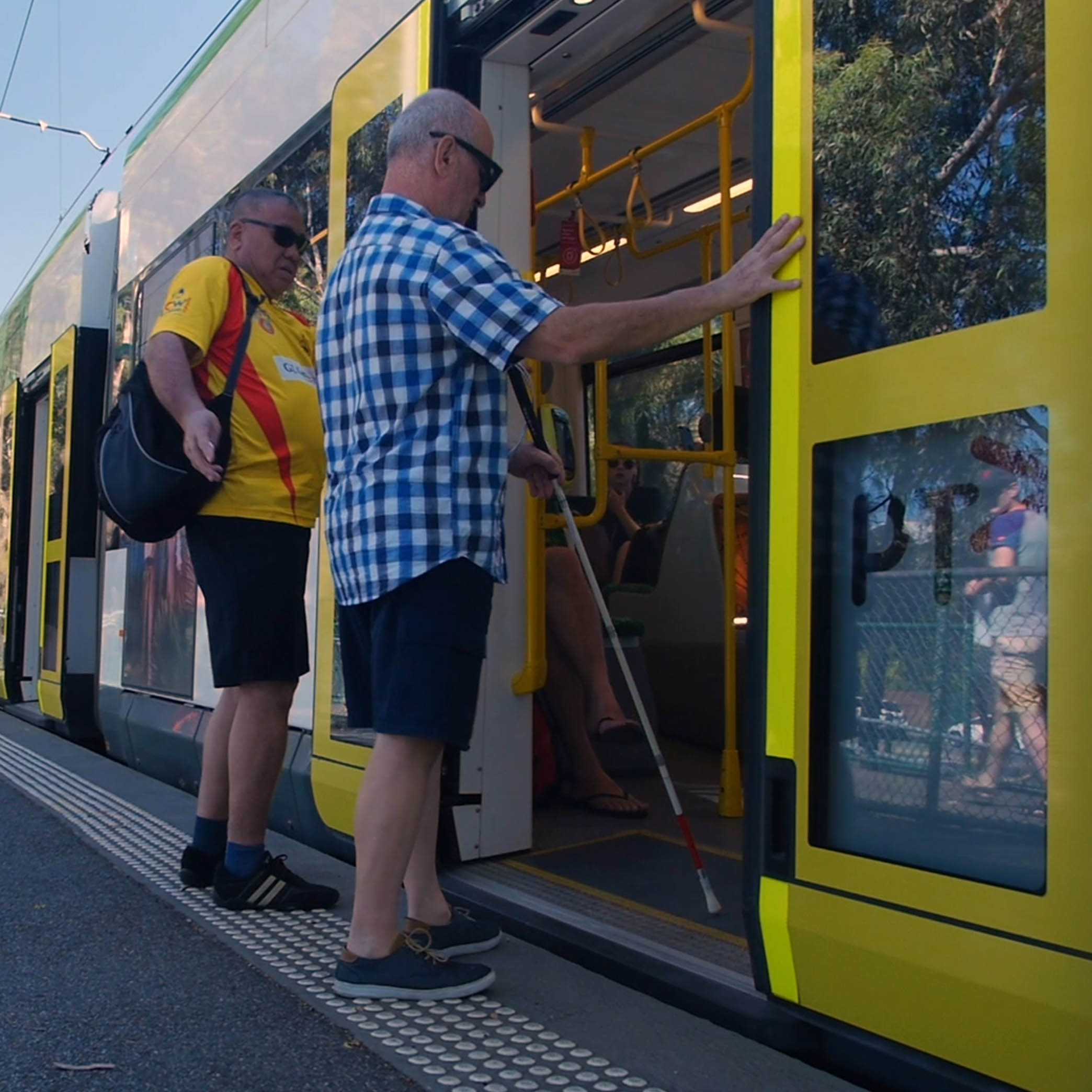 Image shows a white cane user boarding a tram. He is a Caucasian man and is wearing dark blue shorts and a blue and white checked shirt.