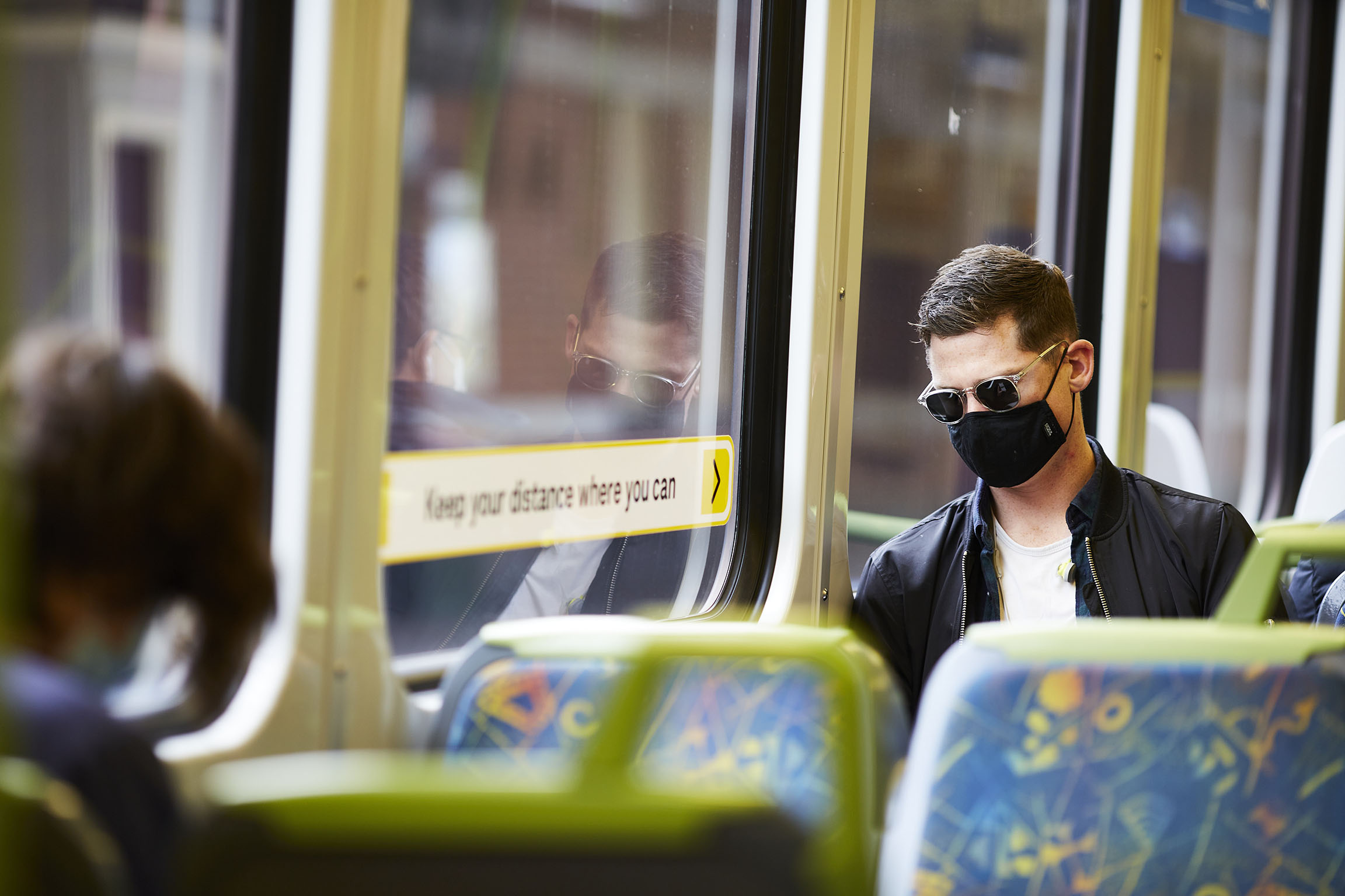 Image is a photograph of a passenger wearing a face mask while travelling on a Metro train. The passenger is a young Caucasian male with short brown hair. He is wearing dark sunglasses with clear frames, a black face mask, black leather jacket and a white t-shirt. He is looking down at his phone and leaning against the window of the train carriage. A blurred station platform is visible through the window.
