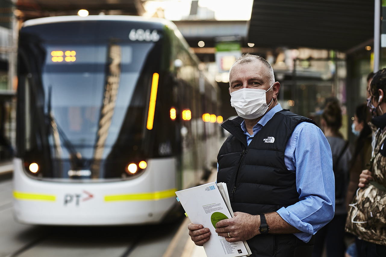 Image is a photograph of Public Transport Ombudsman, Simon McKenzie. Simon is a middle-aged Caucasian man with closely cropped grey hair. He is standing on a tram stop and looking directly at the camera. He is wearing a white face mask, light blue shirt with an open collar and a black puffer vest and is holding some documents in his hands. In the background is a blurred E-Class tram.