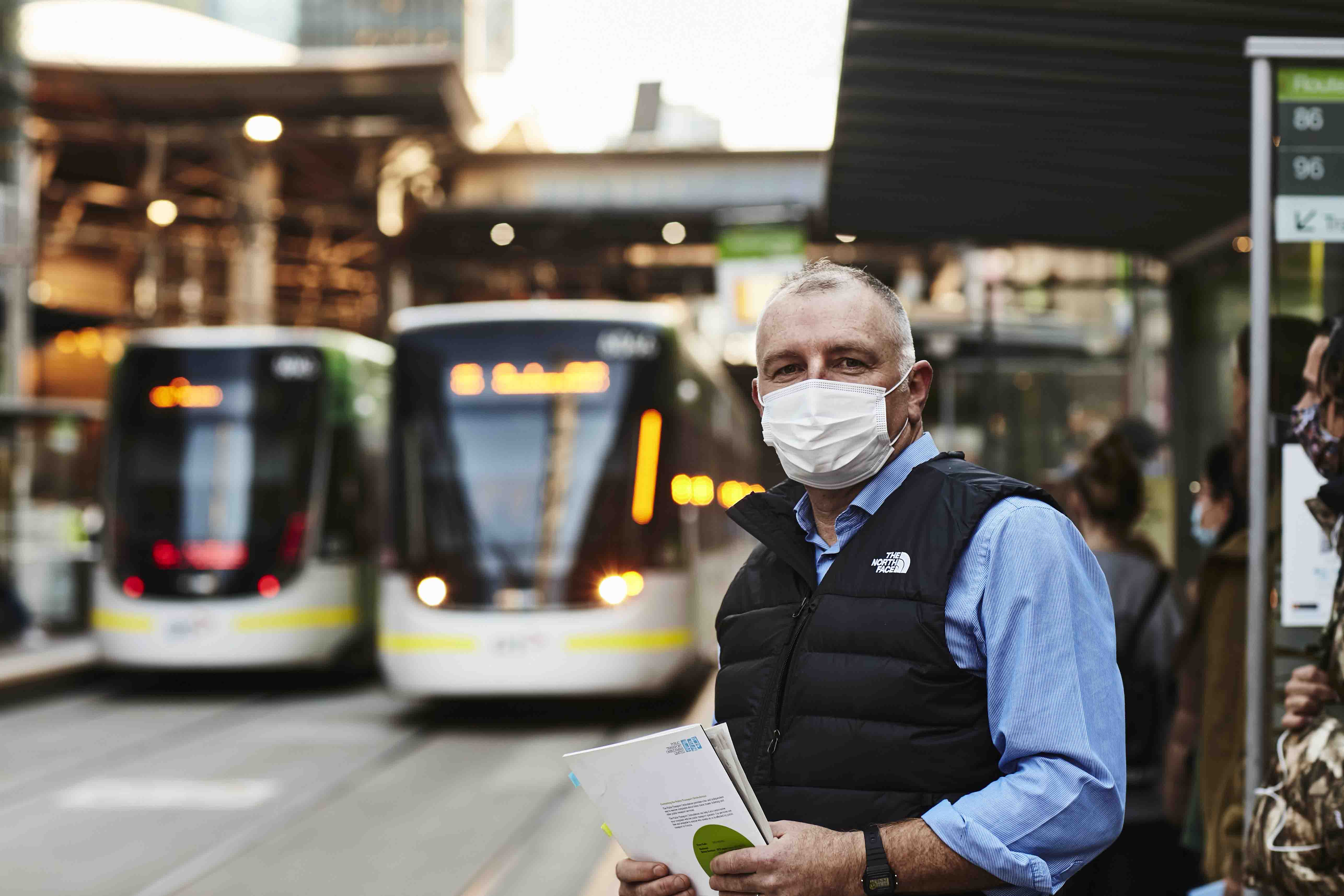 Image is a landscape orientation colour photograph showing Public Transport Ombudsman, Simon McKenzie, waiting at a tram stop in the Melbourne CBD. Simon is a middle-aged Caucasian man with closely cropped grey hair. He is wearing a white facemask and a black puffer vest over a light blue shirt that is open at the collar and rolled-up at the sleeves. He holds some papers in his hands in front of him. In the background and to his left are two trams. The background of the image is blurred from the camera’s depth-of-field.