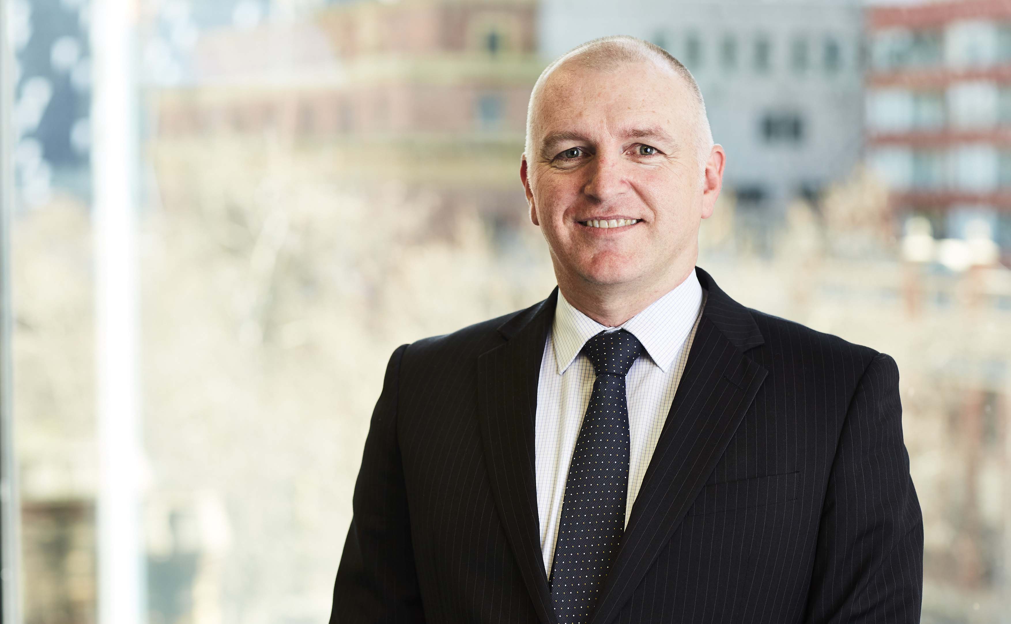 Image shows Public Transport Ombudsman, Simon McKenzie. Simon is a Caucasian man with closely cropped hair and he is smiling for the photo. He is wearing a black pinstripe suit jacket, white checked shirt and navy tie with white dots. The blurred background shows buildings and trees in the Melbourne CBD.