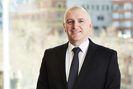 Image shows Public Transport Ombudsman, Simon McKenzie. Simon is a Caucasian man with closely cropped hair and he is smiling for the photo. He is wearing a black pinstripe suit jacket, white checked shirt and navy tie with white dots. The blurred background shows buildings and trees in the Melbourne CBD.