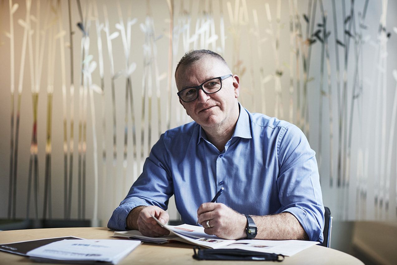 Image shows Public Transport Ombudsman, Simon McKenzie. Simon is a Caucasian man with closely cropped hair. He is wearing a light blue shirt with an open collar and rolled-up sleeves and dark framed glasses. He is sitting at a table with papers and an open magazine in front of him and a pen in his left hand. He is looking directly at the camera. The blurred background shows frosted glass windows with an abstract pattern.
